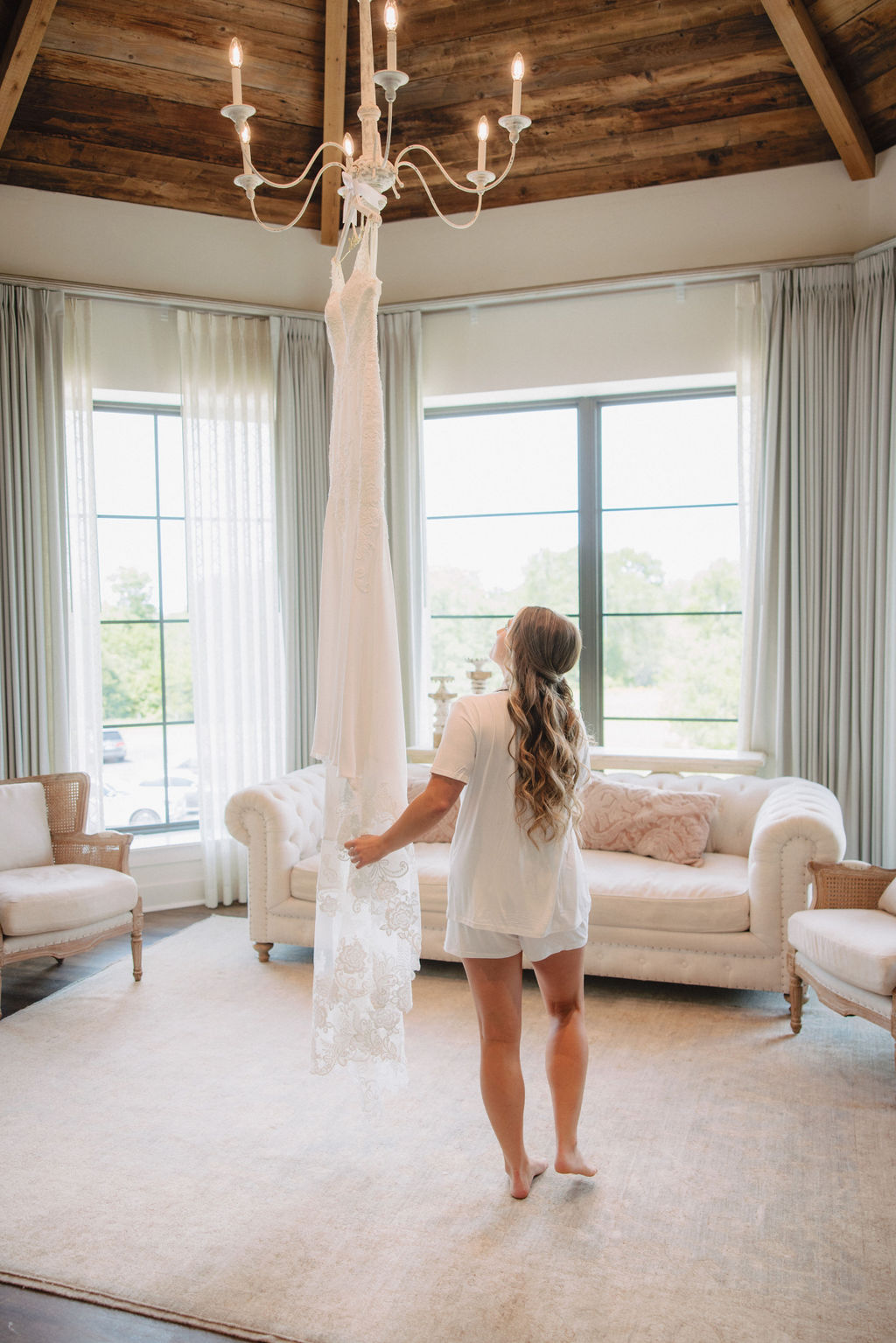 A white wedding dress hangs from a chandelier in a room with a wooden beam ceiling, large windows, and light-colored furniture.