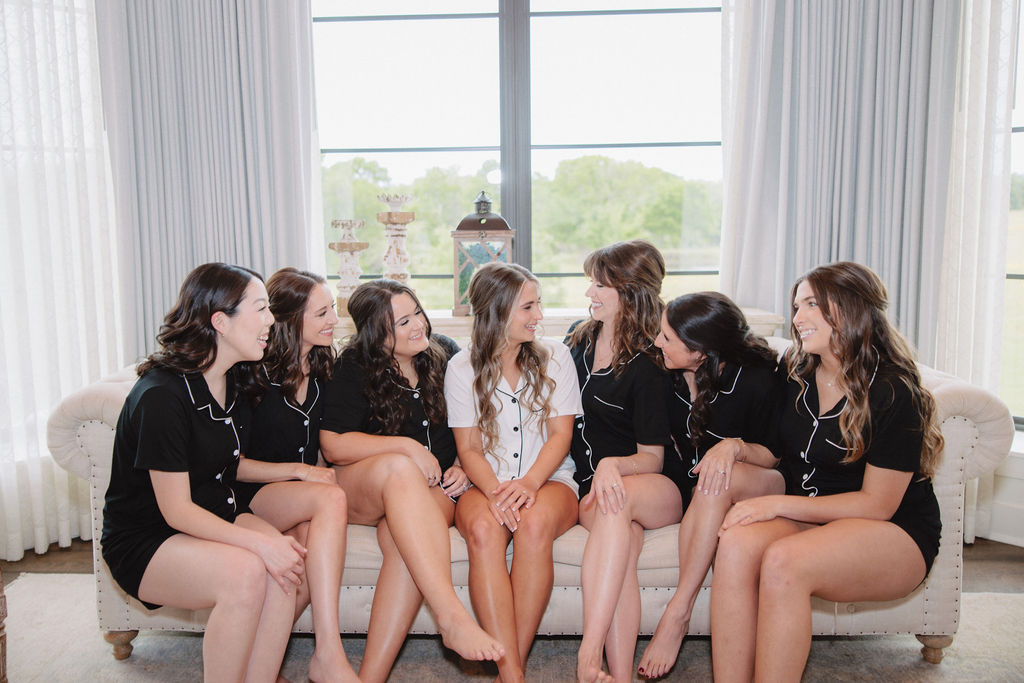 bridesmaid matching black outfits and one person in white sit on a couch, smiling and engaging in conversation at a wedding