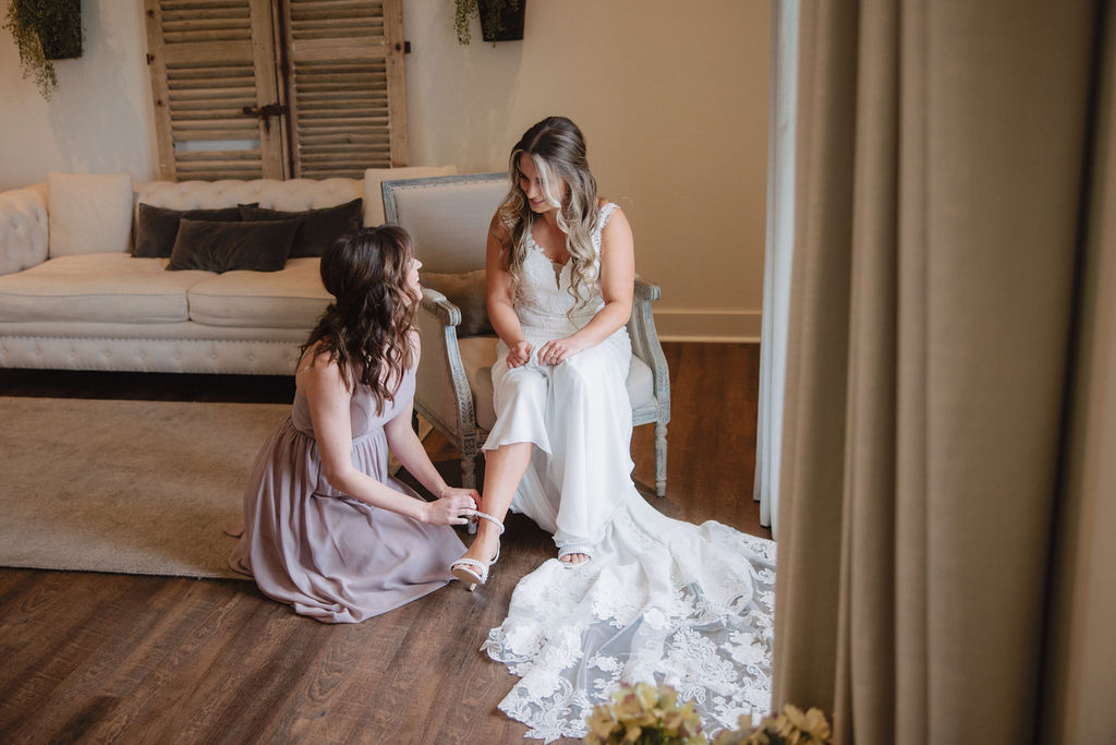 A woman in a blue dress helps another woman adjust her white wedding gown in a well-lit room with large windows and beige furniture.
