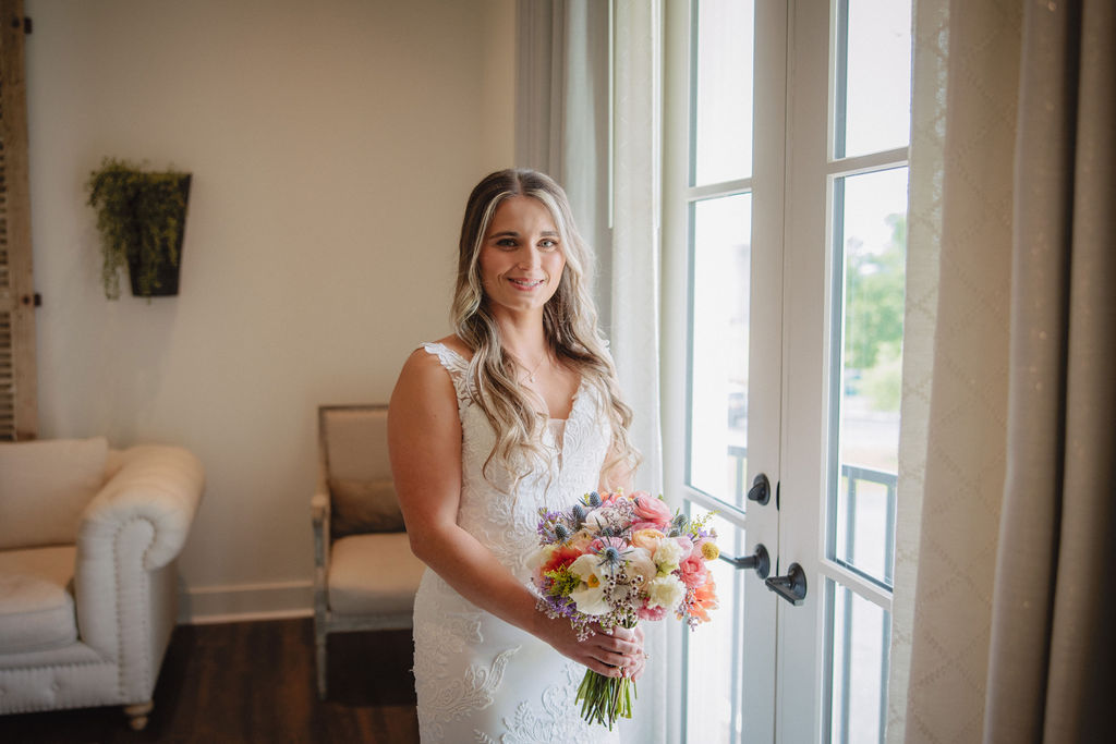 A bride in a white dress holds a colorful bouquet while standing by a glass door inside a well-lit room with a white sofa and wall decor.