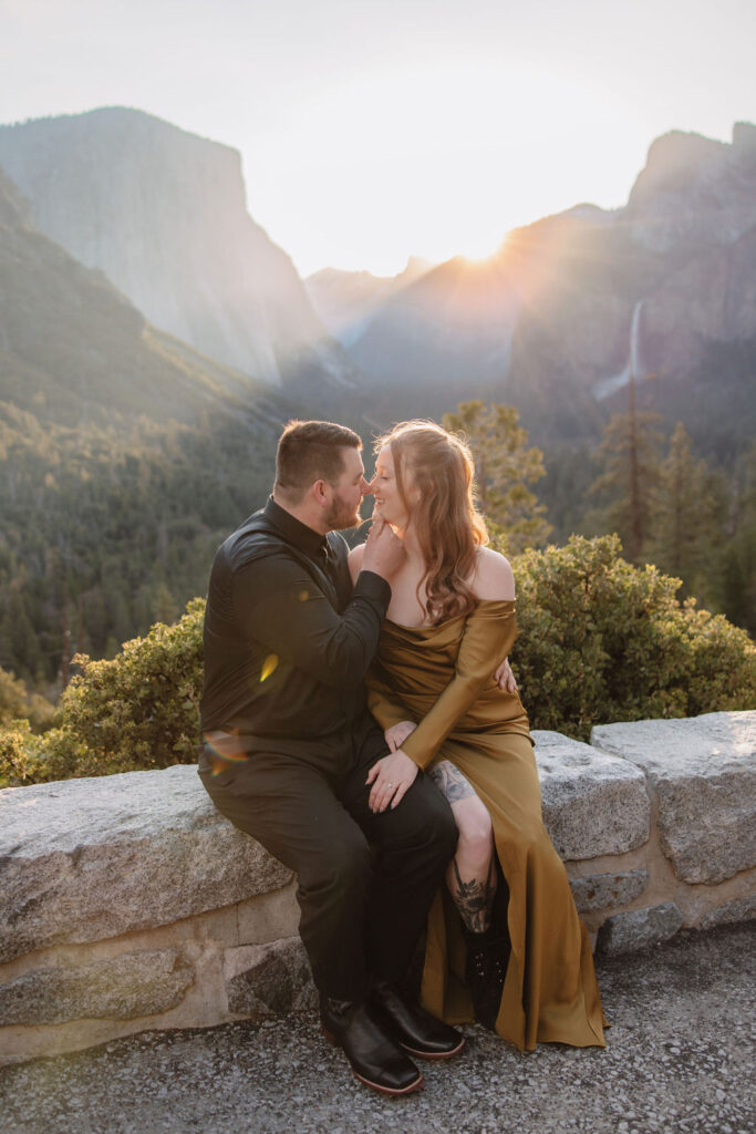 A man lifts a woman in an affectionate pose on a cliffside with a mountainous landscape and the sun setting in the background in Yosemite