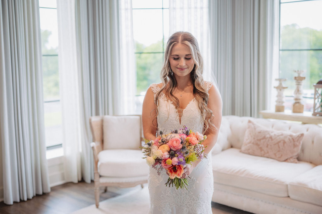 A bride in a white dress holds a colorful bouquet while standing by a glass door inside a well-lit room with a white sofa and wall decor.