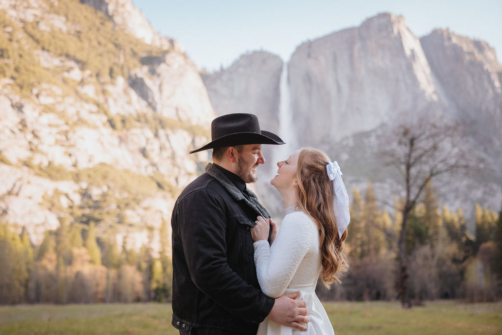 A man in a black hat and a woman in a white dress holding hands in a meadow, with tall misty cliffs in the background at their Yosemite engagement session