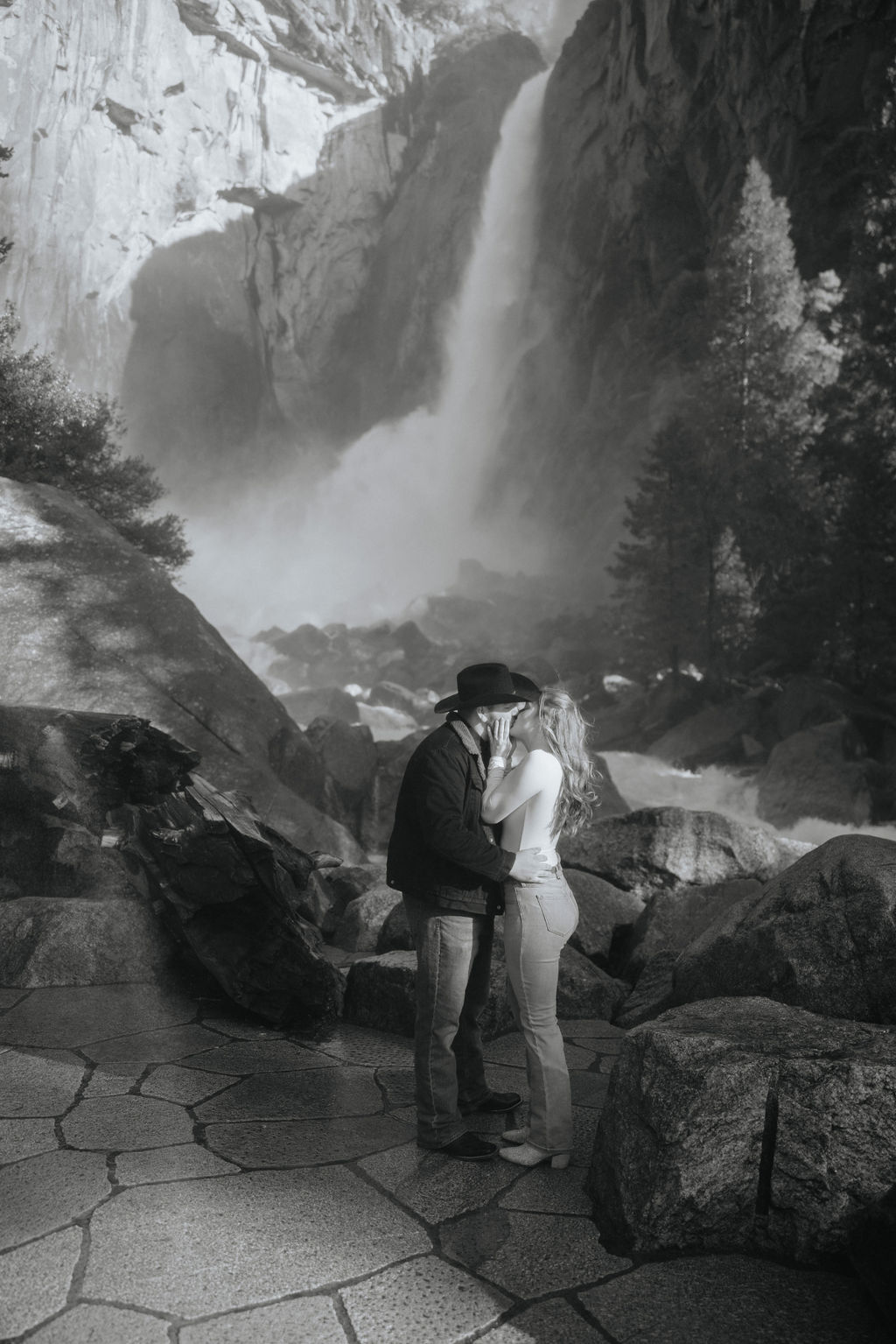 A couple holding hands while walking towards a waterfall in a forest, viewed from behind. the man wears a black hat and the woman a white hat.