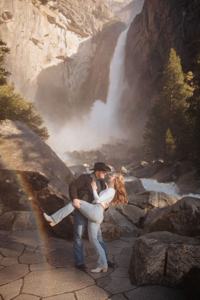 A couple stands close, touching their faces together, in front of a rocky landscape with a large waterfall called Yosemite falls in the background