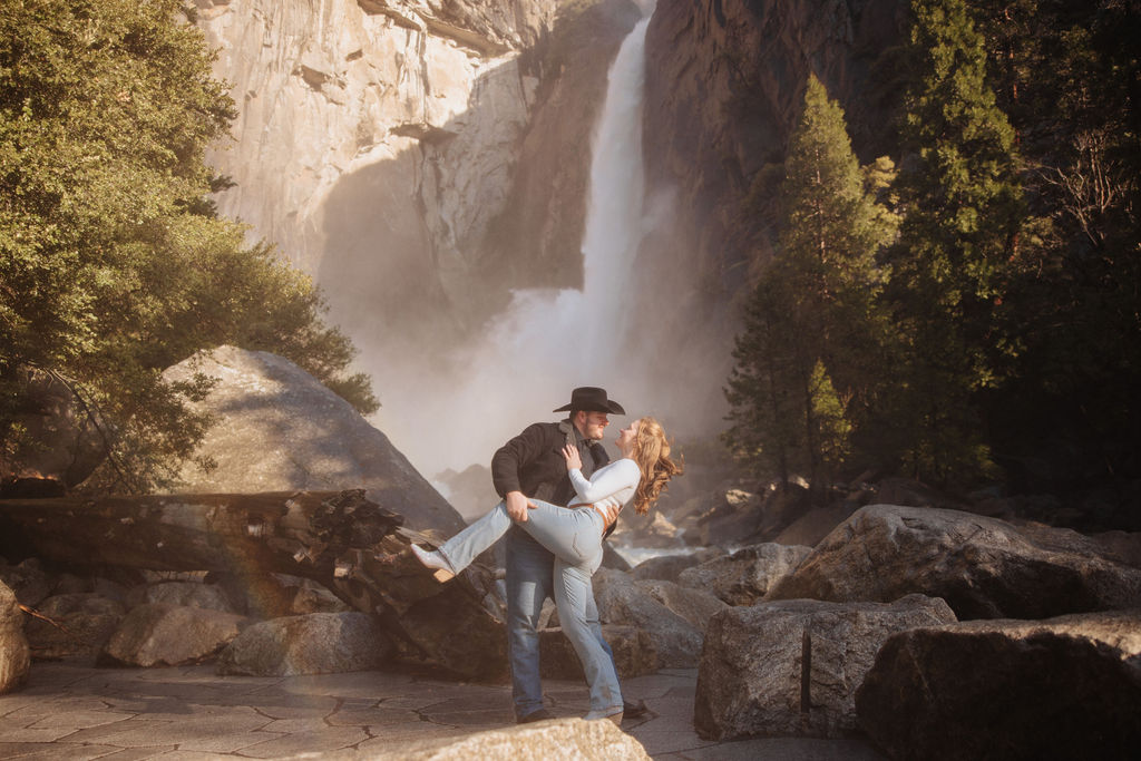 A couple holding hands while walking towards a waterfall in a forest, viewed from behind. the man wears a black hat and the woman a white hat.