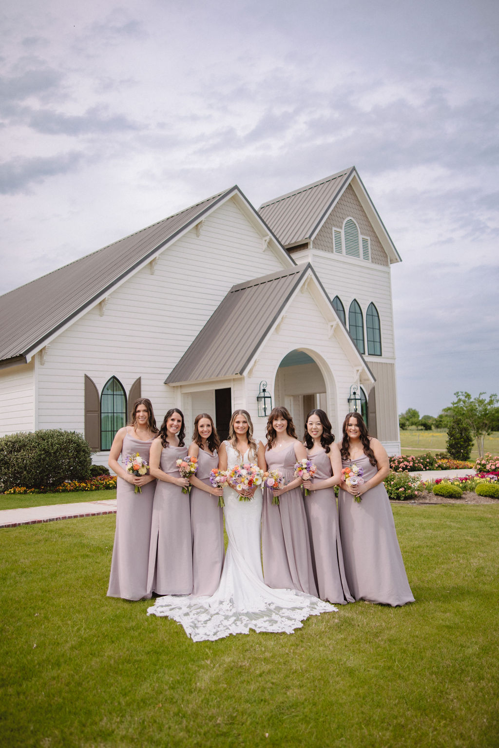 A bride and five bridesmaids in lavender dresses, holding bouquets of flowers, stand together smiling in front of a white chapel on a cloudy day at deep in the heart farms