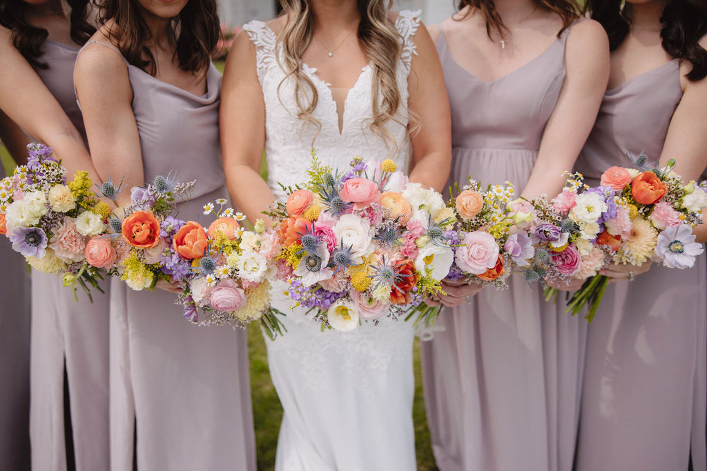 A bride and five bridesmaids in lavender dresses, holding bouquets of flowers, stand together smiling in front of a white chapel on a cloudy day at deep in the heart farms