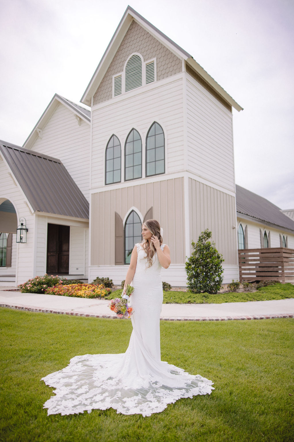 A bride in a white wedding dress holding a bouquet stands on a lawn in front of a tall, white, church-like building with pointed arched windows at deep in the heart farms