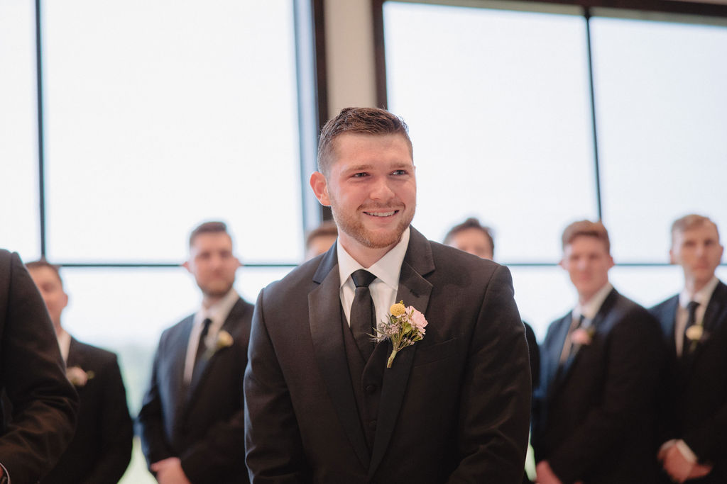 A groom stands at the altar with his groomsmen lined up behind him in a church with large windows. An officiant in a black suit stands nearby holding a book.