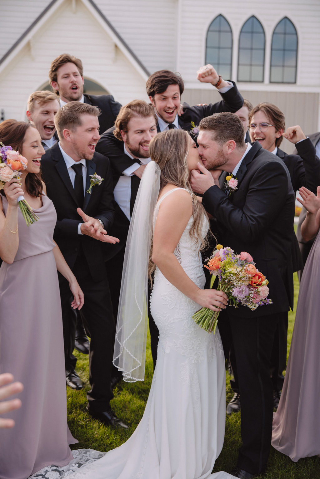 A bride and groom stand surrounded by their wedding party outside a white chapel, with the group cheering and smiling. The wedding party members are wearing matching dresses and suits at deep in the heart farms wedding venue
