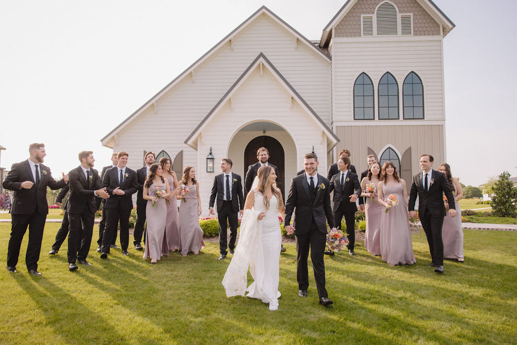 A bride and groom stand surrounded by their wedding party outside a white chapel, with the group cheering and smiling. The wedding party members are wearing matching dresses and suits at deep in the heart farms wedding venue