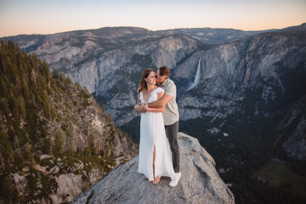 Couple standing on a rock with yosemite falls in the background holding each other during their engagement photoshoot at glacier point in yosemite. 