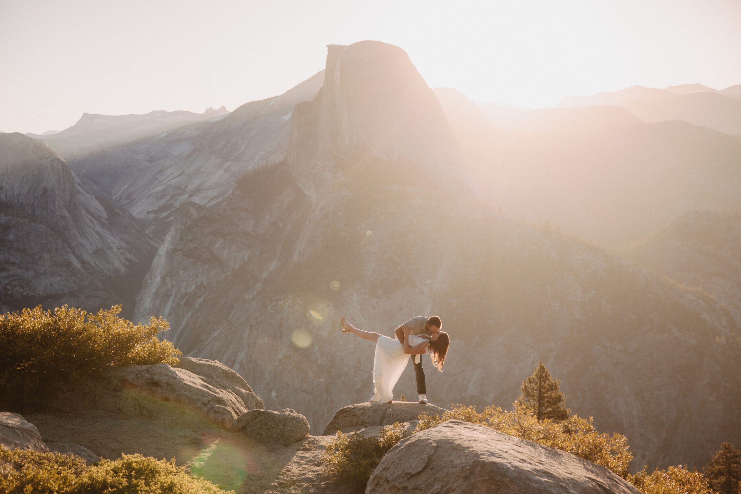 A couple stands on a cliff at sunset, sharing a kiss with a mountainous landscape behind them.