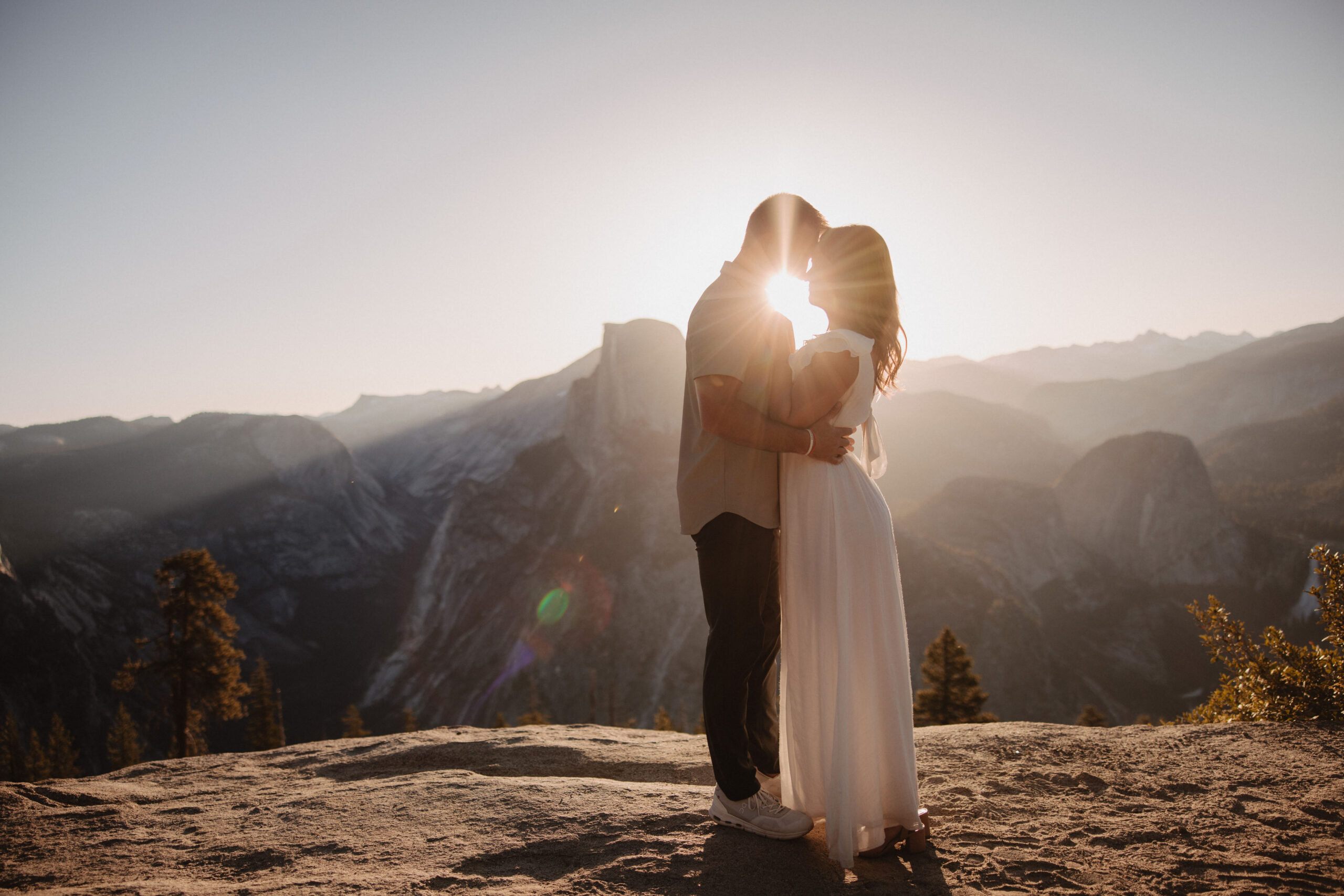 A couple stands on a cliff at sunset, sharing a kiss with a mountainous landscape behind them.