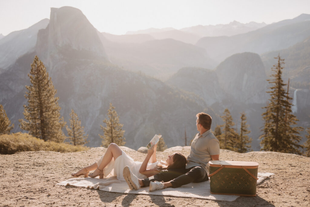 Couple has a picnic at glacier point. Woman reads book at sunrise for their engagement photoshoot