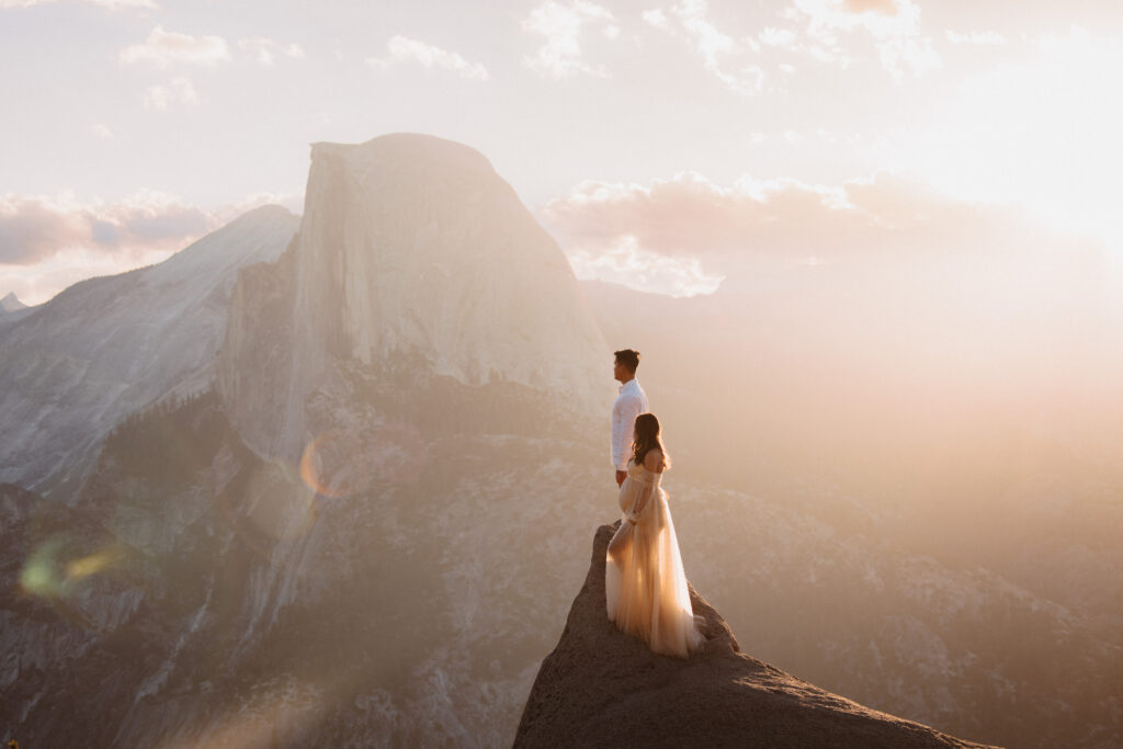 Couple standing on rock looking out at glacier point in yosemite for their maternity photoshoot at sunrise. 