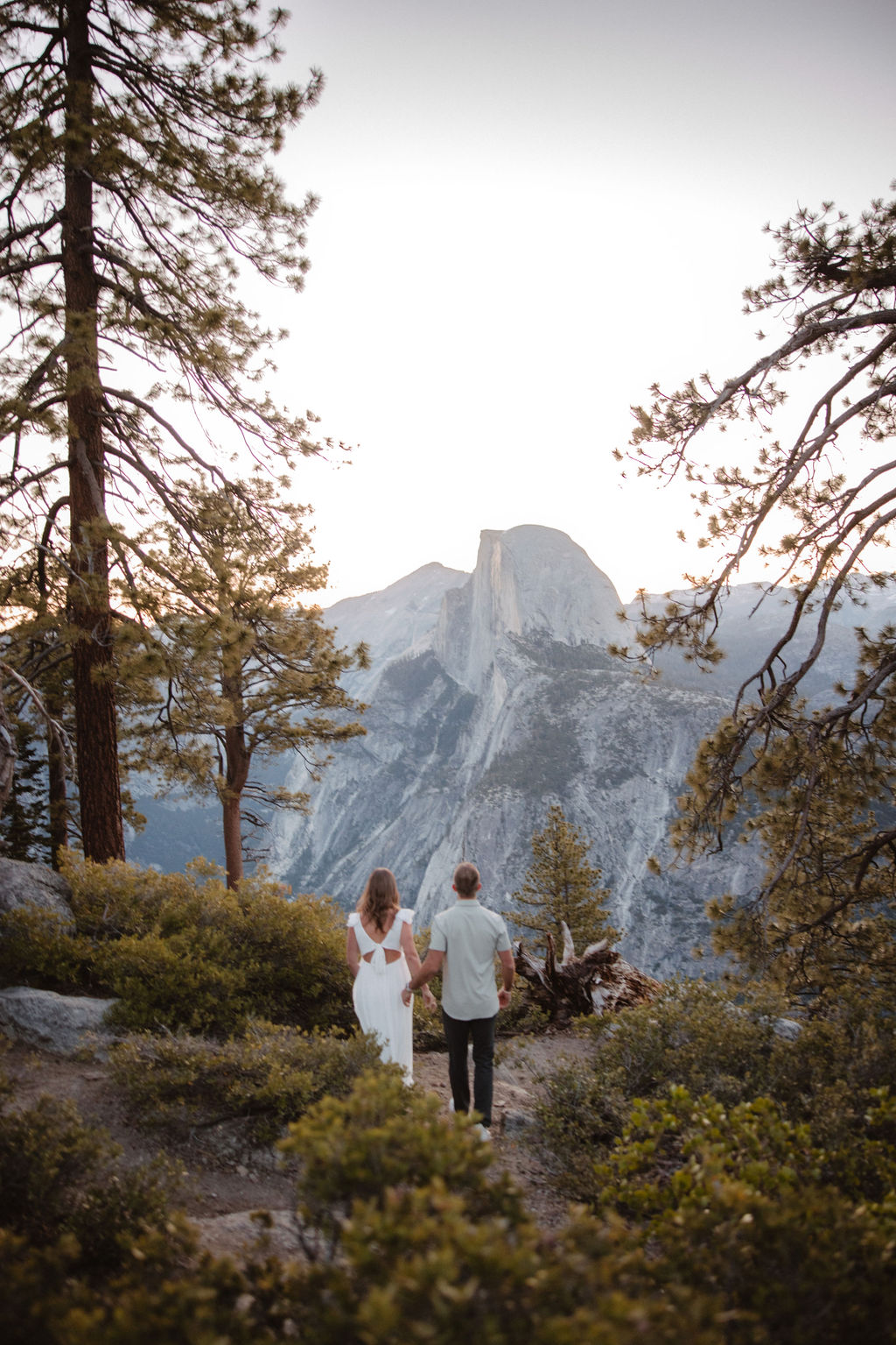 A couple stands embracing amidst trees, with a mountain range featuring a prominent rock formation in the background during their Glacier Point engagement photos