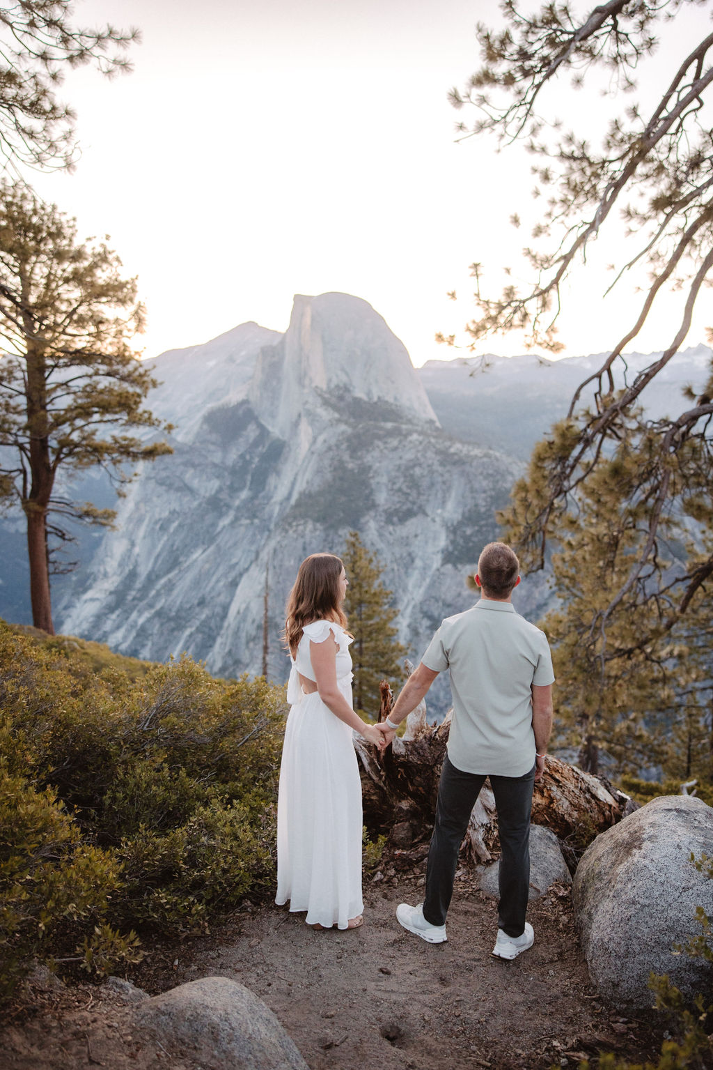 A couple stands embracing amidst trees, with a mountain range featuring a prominent rock formation in the background during their Glacier Point engagement photos