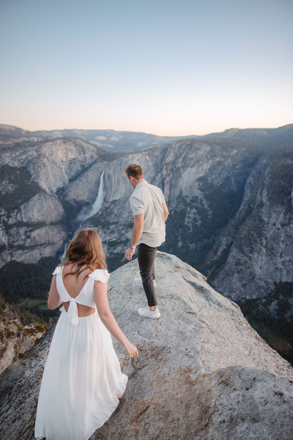 A couple stands on a rocky ledge overlooking a forested mountainous landscape, with the man embracing the woman from behind.