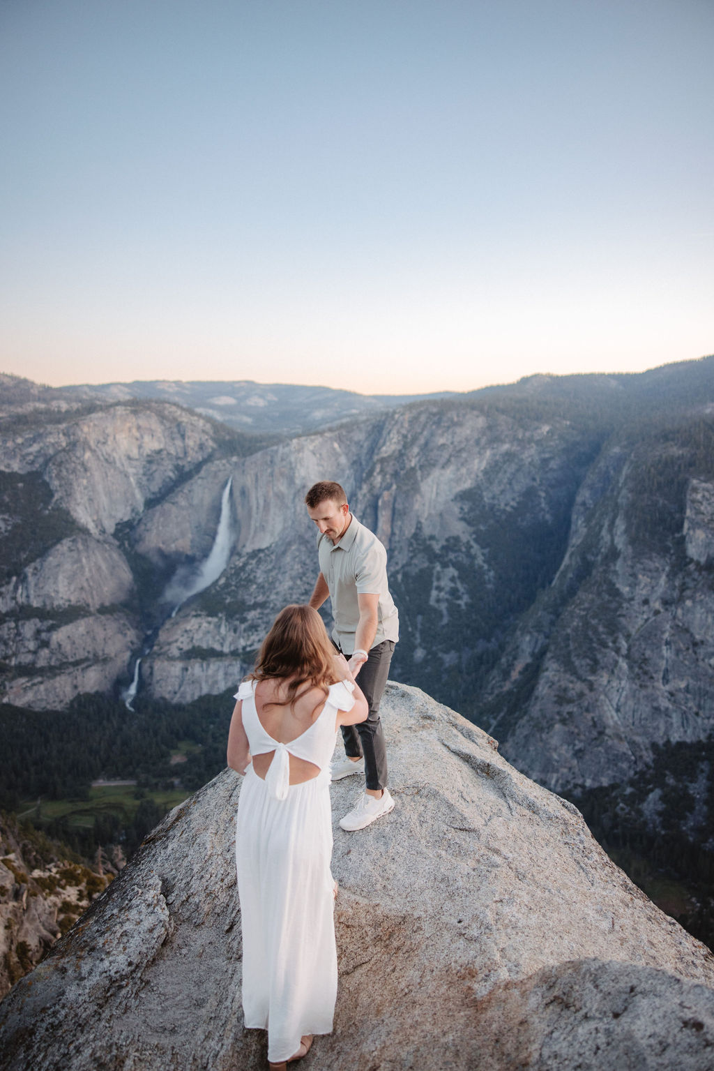 A couple stands on a rocky ledge overlooking a forested mountainous landscape, with the man embracing the woman from behind.