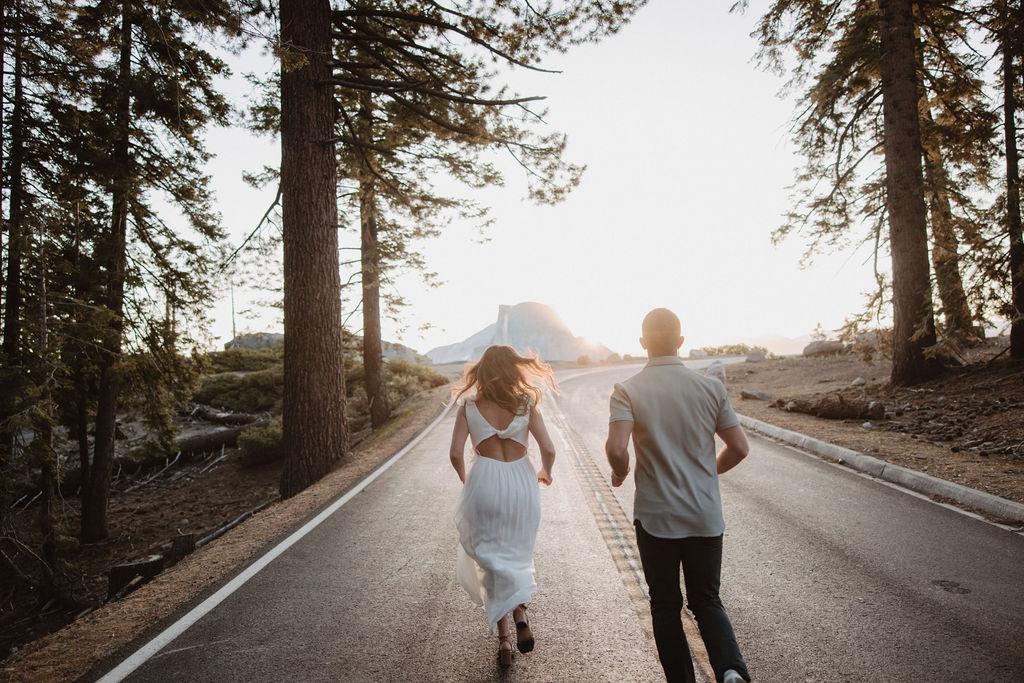 A man and a woman run down a rural, tree-lined road at sunset, with mountains visible in the background during their glacier point engagement photos