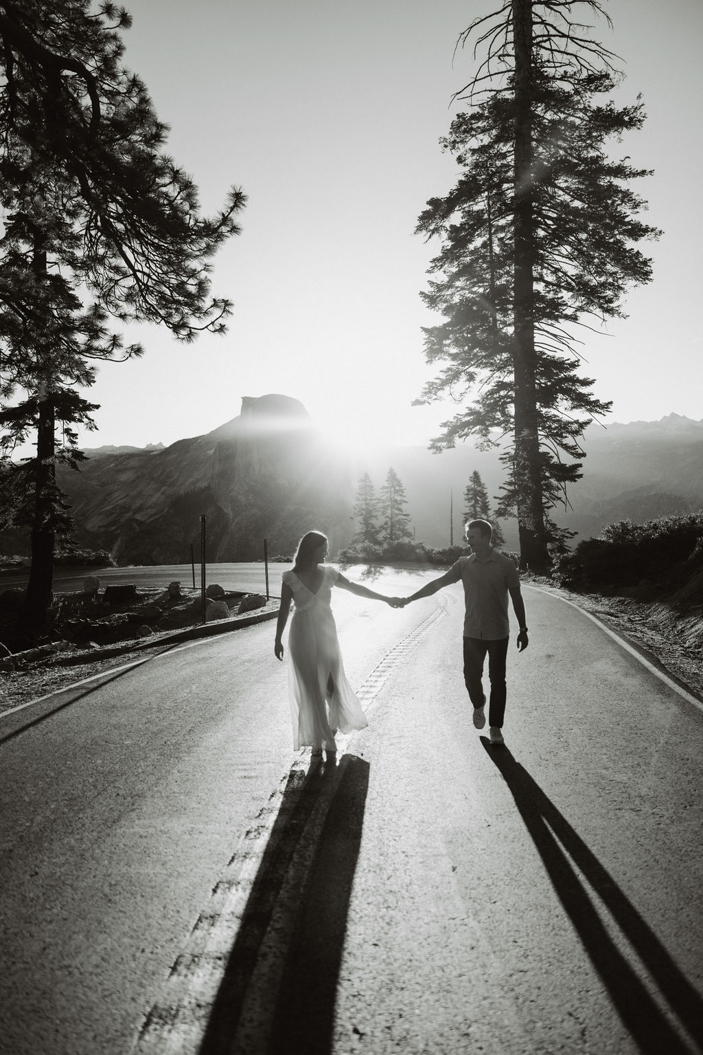 A man and a woman run down a rural, tree-lined road at sunset, with mountains visible in the background during their glacier point engagement photos