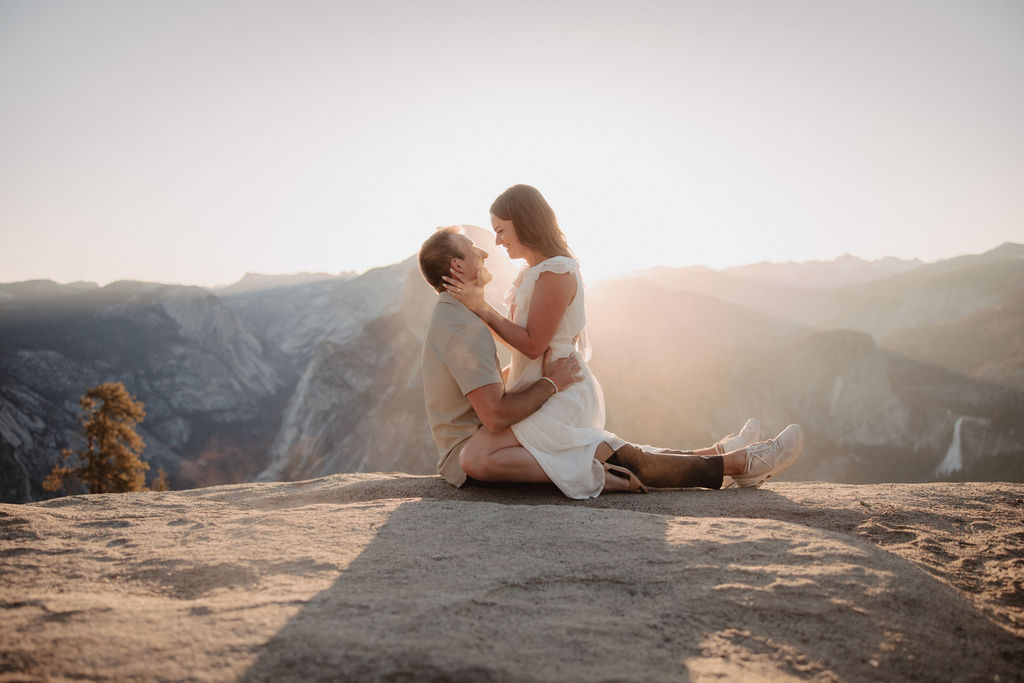 A man lifts a woman in a white dress as they embrace against a backdrop of a mountainous landscape with a setting sun during their glacier point engagement photos