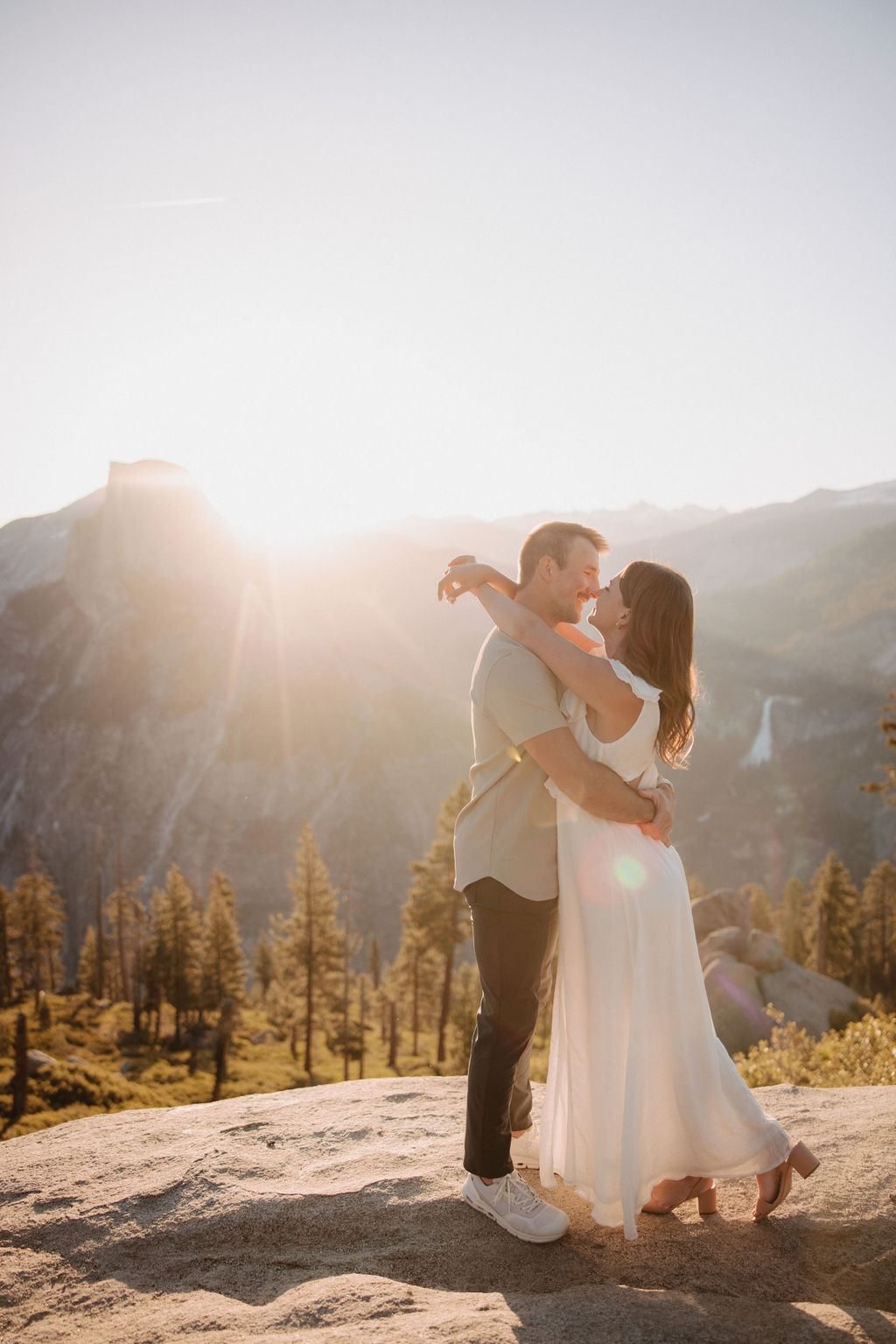 A man lifts a woman in a white dress as they embrace against a backdrop of a mountainous landscape with a setting sun during their glacier point engagement photos