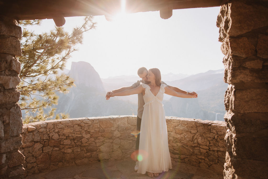 A stone cabin with a wooden roof is situated on a mountainside. Three people stand on the stone steps, with a large mountain visible in the hazy background for their glacier point engagement photos