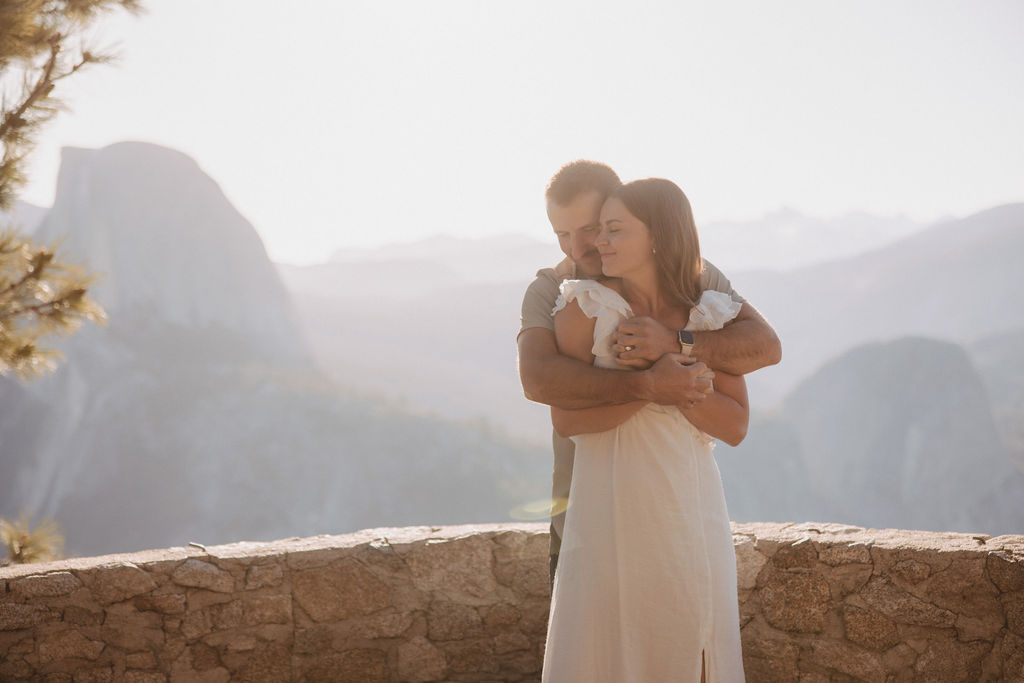 A stone cabin with a wooden roof is situated on a mountainside. Three people stand on the stone steps, with a large mountain visible in the hazy background for their glacier point engagement photos