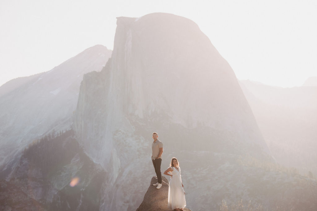 Two people stand on a rock ledge, with a large, hazy mountain in the background. One is dressed in light-colored suit and pants, the other in a white dress. The scene is backlit by the sun.