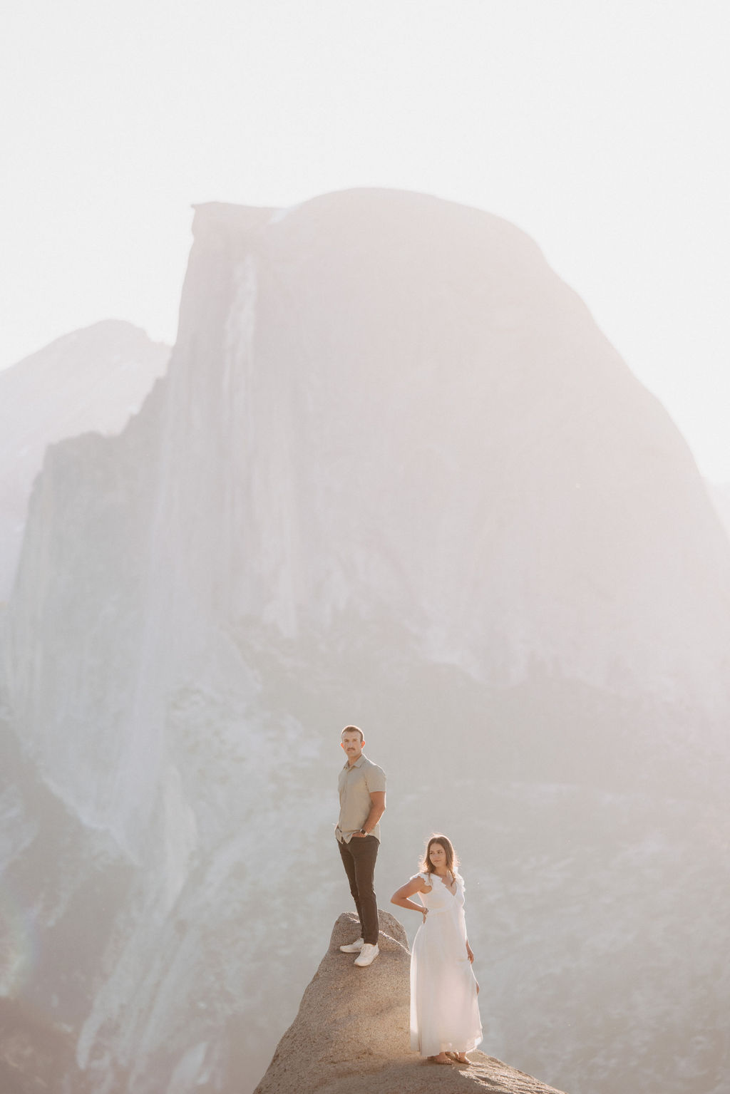Two people stand on a rock ledge, with a large, hazy mountain in the background. One is dressed in light-colored suit and pants, the other in a white dress. The scene is backlit by the sun.