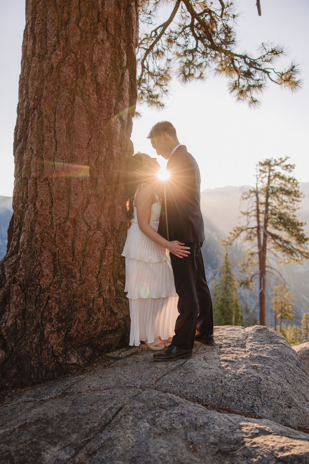 A couple stands close to each other in front of a large tree with sunlight filtering through the branches and distant mountains in the background for their Yosemite Elopement 