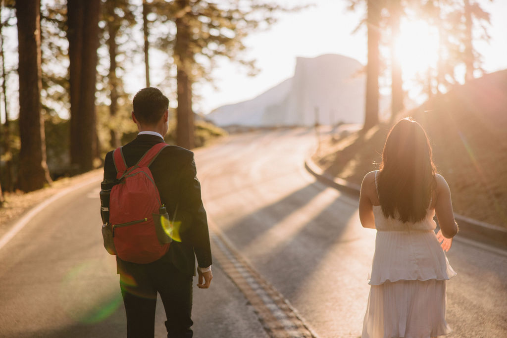 Two people holding hands while walking on a tree-lined road at sunrise or sunset, with mountains visible in the background at their Yosemite elopement 