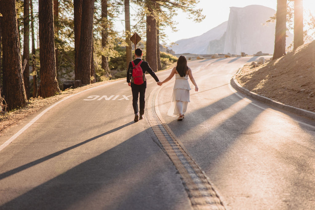 Two people holding hands while walking on a tree-lined road at sunrise or sunset, with mountains visible in the background at their Yosemite elopement 