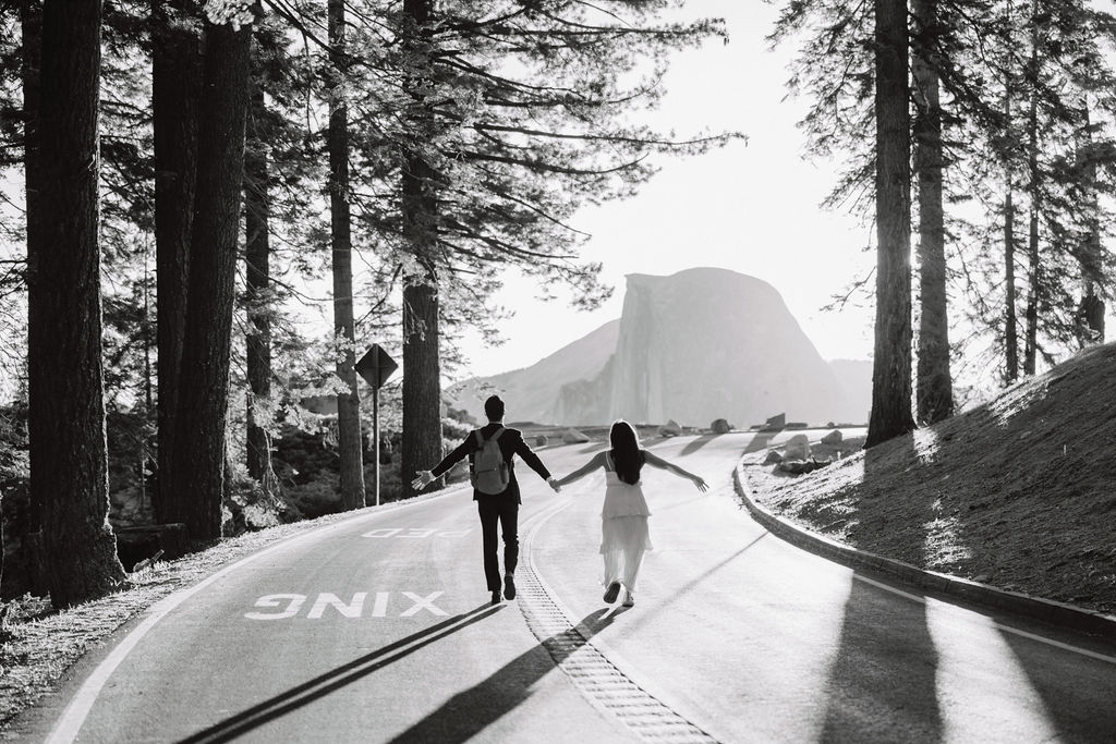 Two people holding hands while walking on a tree-lined road at sunrise or sunset, with mountains visible in the background at their Yosemite elopement 