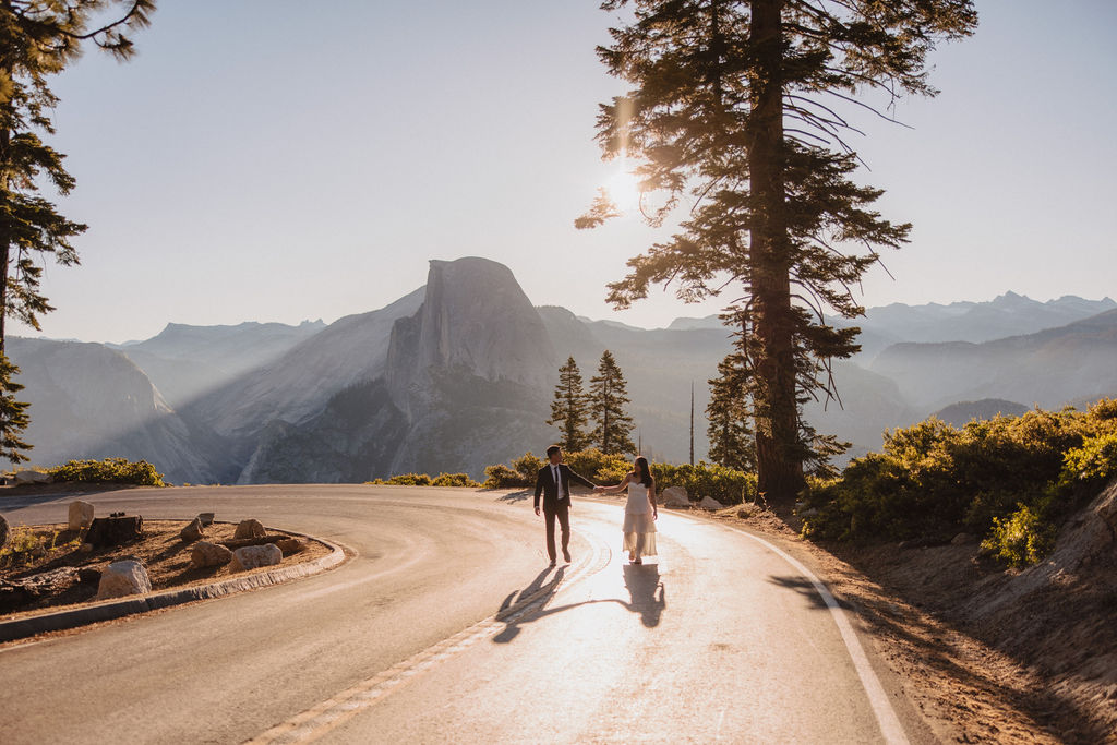 Two individuals walk down a winding road at sunset with mountainous scenery and trees in the background. The sun is partially visible behind the trees, casting shadows on the road at their Yosemite elopement 