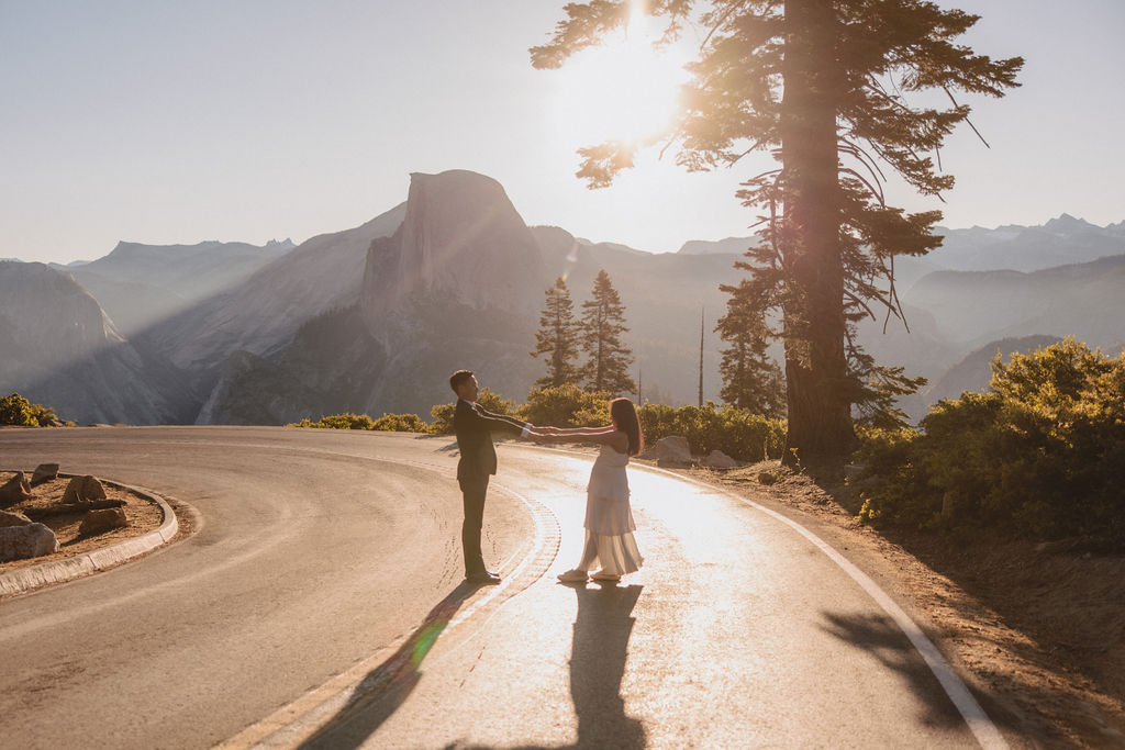 Two individuals walk down a winding road at sunset with mountainous scenery and trees in the background. The sun is partially visible behind the trees, casting shadows on the road at their Yosemite elopement 