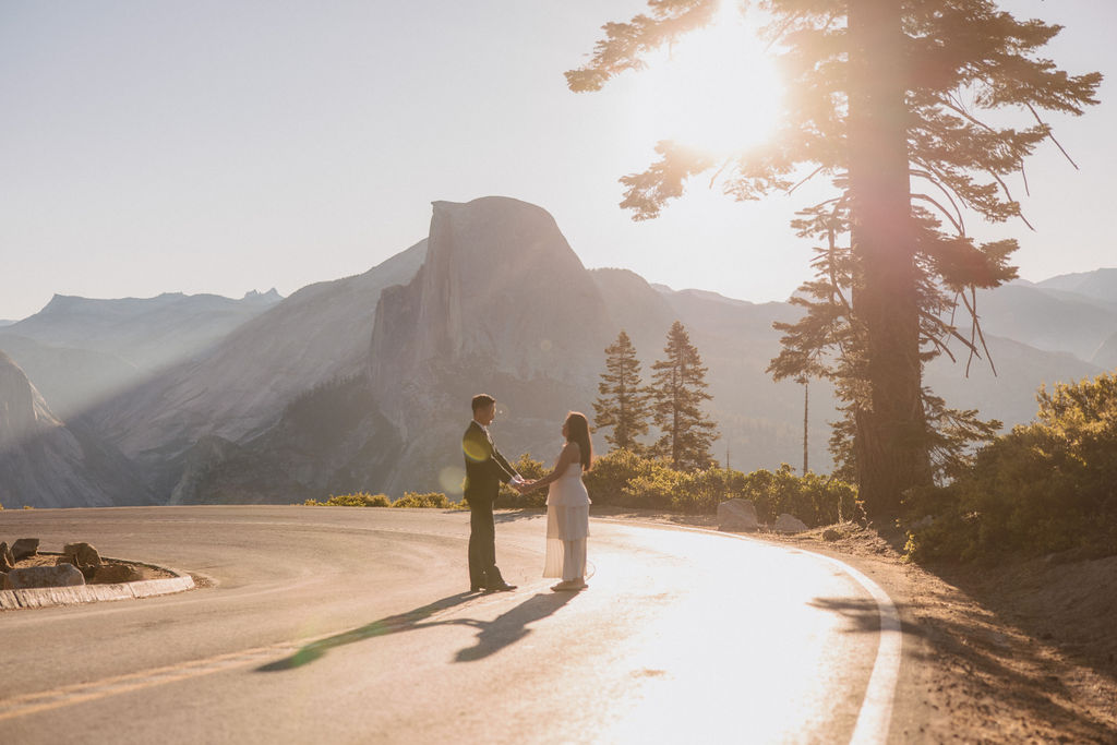 Two people holding hands while walking on a tree-lined road at sunrise or sunset, with mountains visible in the background at their Yosemite elopement 