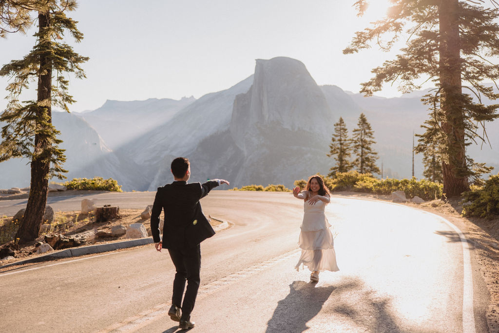 Two people holding hands while walking on a tree-lined road at sunrise or sunset, with mountains visible in the background at their Yosemite elopement 