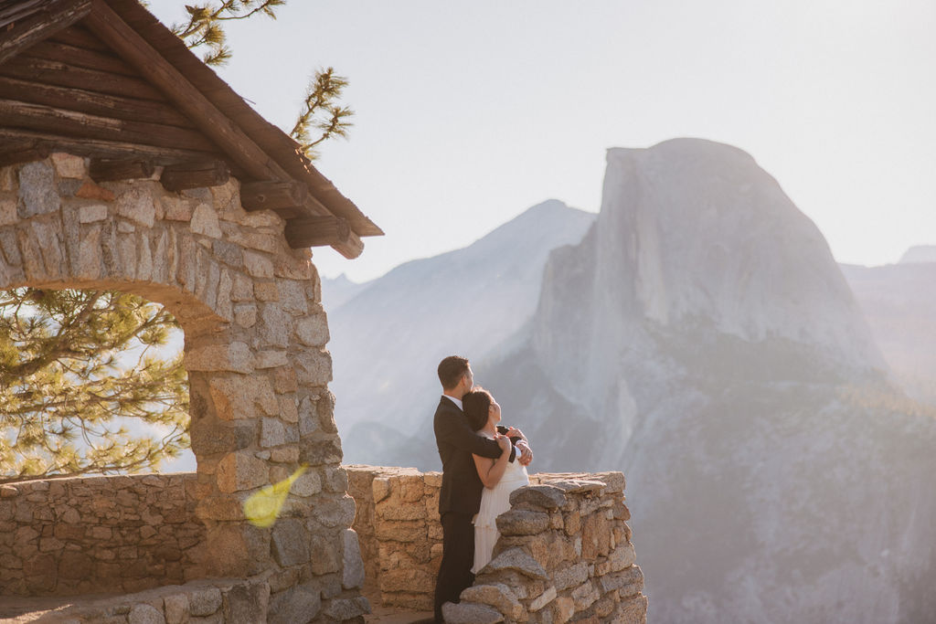 A couple in wedding attire embraces on a stone terrace with a wooden roof, overlooking a mountainous landscape illuminated by sunlight for their Yosemite elopement 