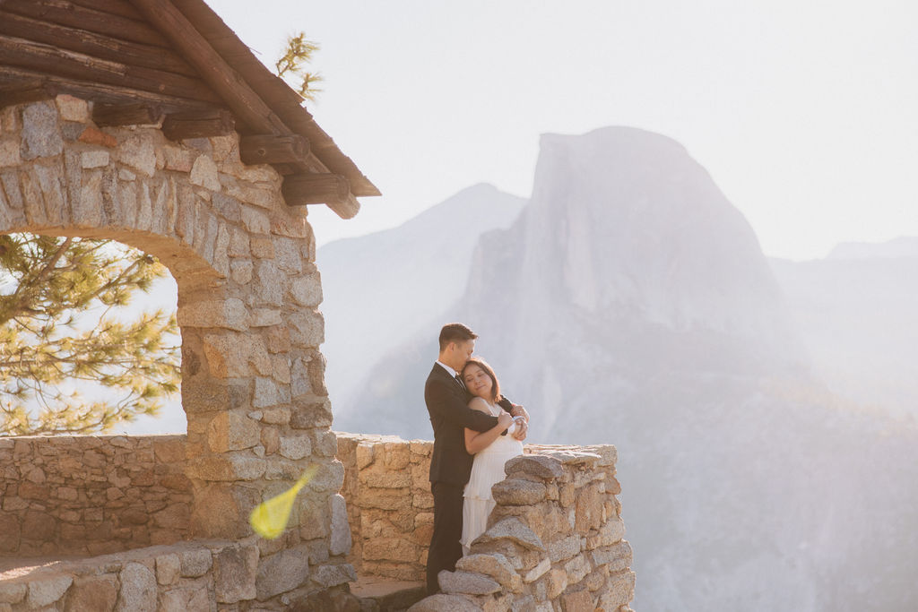 A couple in wedding attire embraces on a stone terrace with a wooden roof, overlooking a mountainous landscape illuminated by sunlight for their Yosemite elopement 