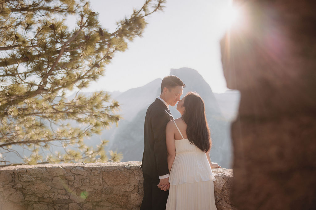 A couple in wedding attire embraces on a stone terrace with a wooden roof, overlooking a mountainous landscape illuminated by sunlight for their Yosemite elopement 