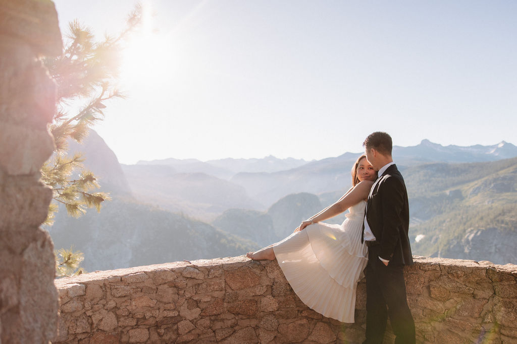 A couple in wedding attire embraces on a stone terrace with a wooden roof, overlooking a mountainous landscape illuminated by sunlight for their Yosemite elopement 