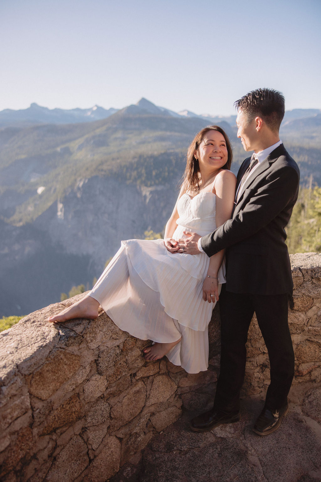 A couple in wedding attire embraces on a stone terrace with a wooden roof, overlooking a mountainous landscape illuminated by sunlight for their Yosemite elopement 