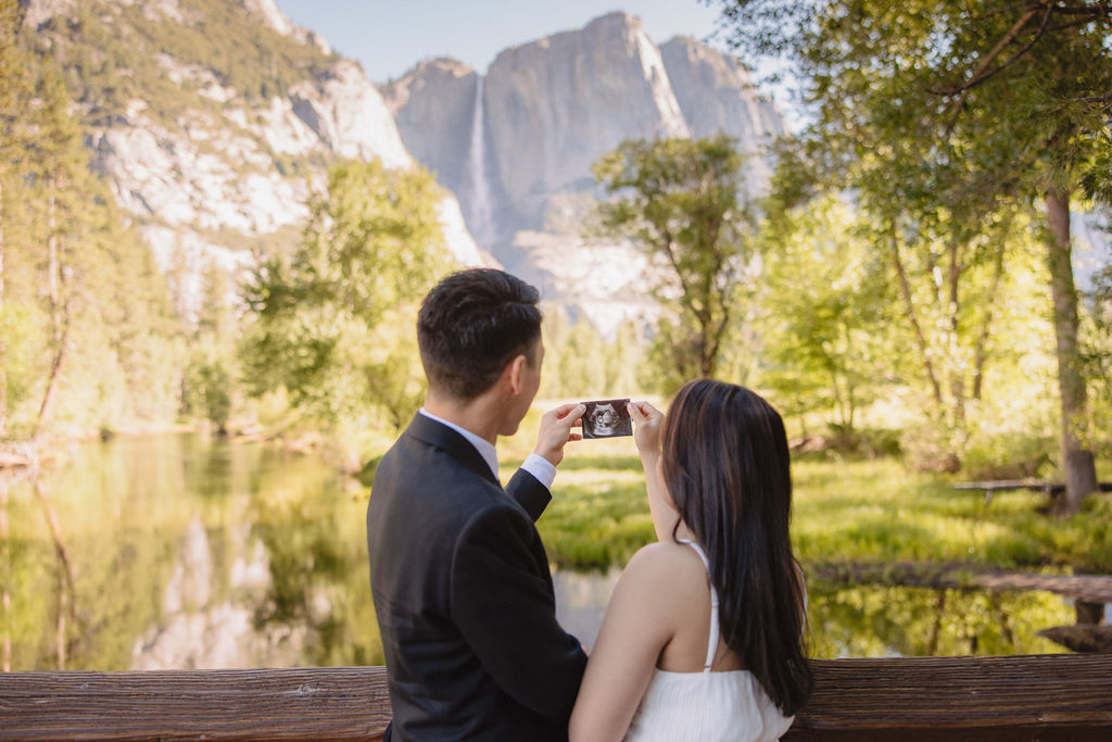 A couple stands on a bridge, with their backs to the camera, taking a photo of a scenic mountain and river landscape at their Yosemite elopement 