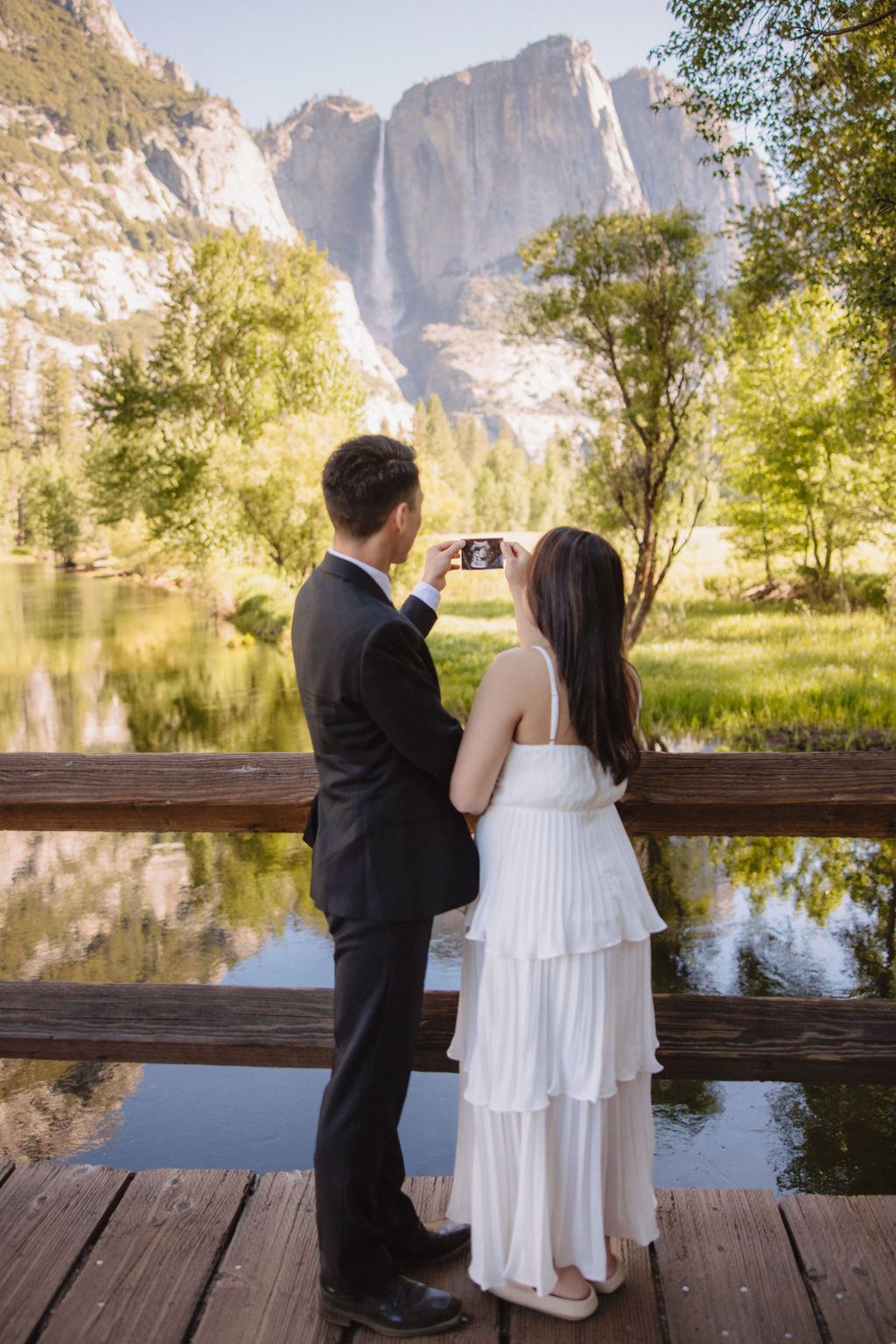 A couple stands on a bridge, with their backs to the camera, taking a photo of a scenic mountain and river landscape at their Yosemite elopement 