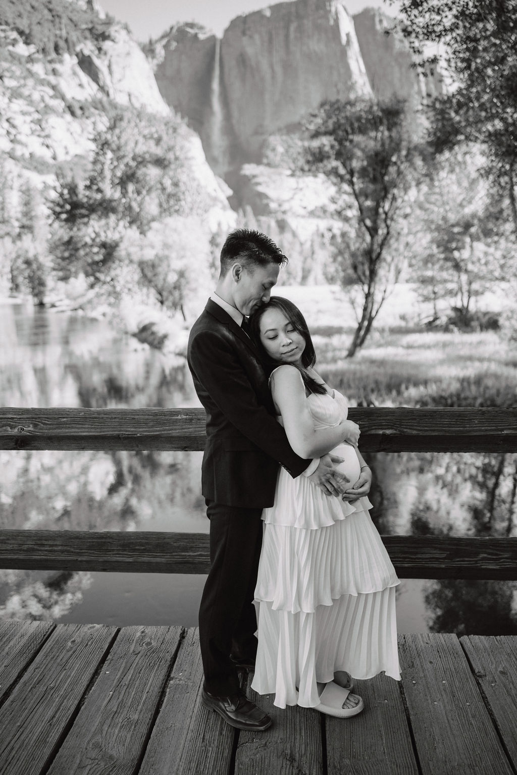 A couple stands on a bridge, with their backs to the camera, taking a photo of a scenic mountain and river landscape at their Yosemite elopement 