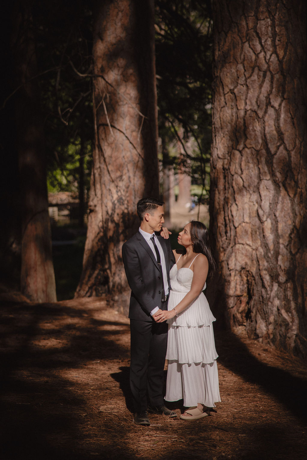 A person in a white dress stands against a large tree trunk, with one hand on a hat, in a forest setting.